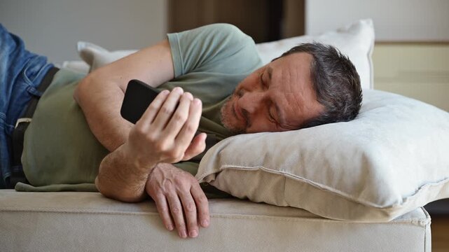 Lazy couch potato guy using mobile phone while laying on living room sofa, 4K