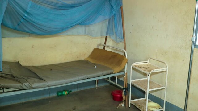 Empty bed with a blue mosquito net and rusty shelves in a poorly maintained hospital ward in Cameroon. Static wide shot.
