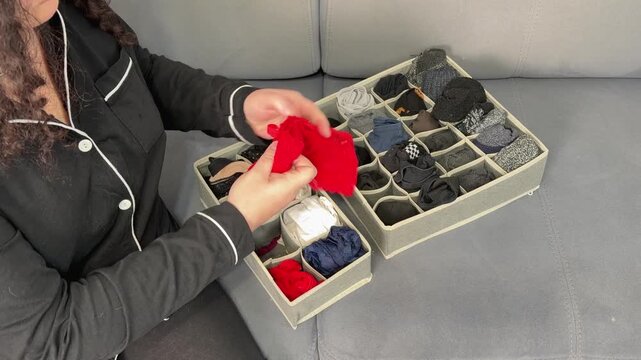 woman, organizing, socks, home, tidying, folding, A woman in pajamas sits on sofa neatly folds socks, placing them into compartmentalized fabric organizers. Home organization, decluttering, tidying up