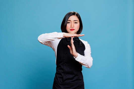 Confident asian waitress dressed in restaurant uniform making timeout gesture with arms portrait. Woman receptionist showing take a break sign with hands and looking at camera with serious expression