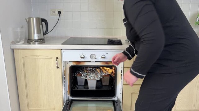 A woman with dark curly hair takes freshly baked loaves of bread out of a hot oven. She uses an oven mitt for protection and places the hot bread on the kitchen counter to cool. Domestic life scene