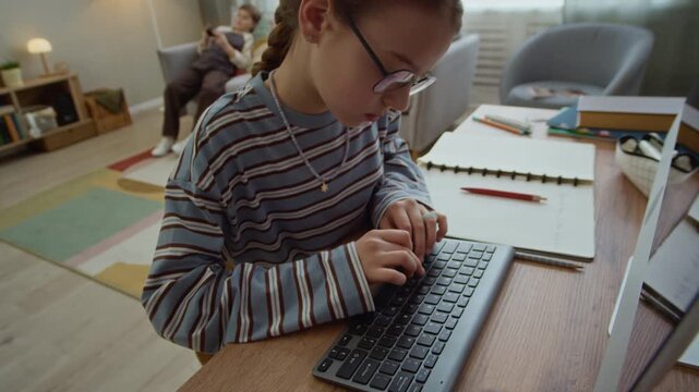 Handheld shot of focused gen Alpha Caucasian girl using computer while doing homework in living room with brother playing console in background