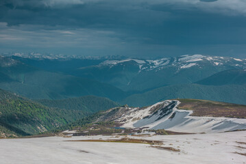 Dramatic landscape with small lake among snowfields, thawed ground, forest and snow cornice on long spur with view to big snowy mountain range far away under cloudy sky. Sunlight and shadows of clouds © Daniil