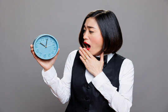 Tired overslept asian waitress in uniform holding circle dial alarm clock and checking time. Restaurant sleepy woman receptionist looking at round retro watch while yawning