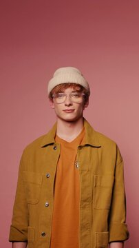 Calm young man with glasses and a beanie looks at the camera. He is wearing an orange t-shirt and a yellow jacket. Vertical, medium shot.