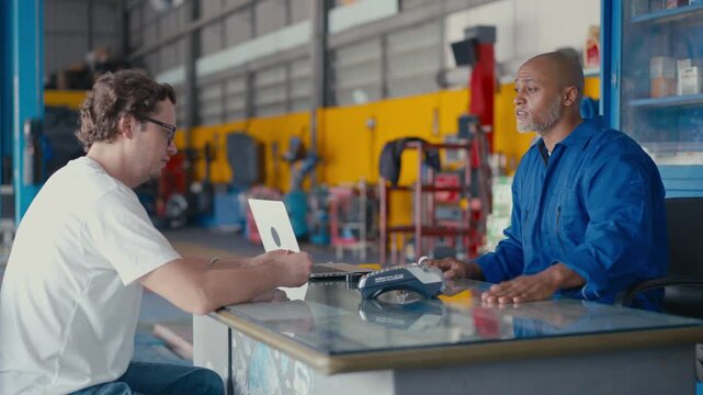 Mechanic and customer discuss repair service in industrial workshop, meeting at garage table with laptop and tool, conversation at workshop desk for SEO friendly title