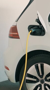 A sleek electric vehicle with a clean white exterior pulls up to a charging station under soft indoor lighting, its port connected by a yellow charging cable.