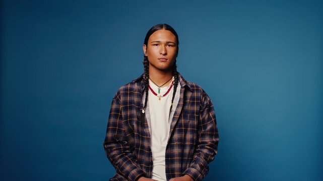 Young Indigenous man with braids and traditional necklaces sits against a vibrant blue backdrop, conveying a sense of pride and cultural identity in a medium close-up shot.