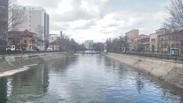 A wide view of a river flowing through a city. Buildings and trees line the banks. The clouds above create changing light. People walk on the paths nearby.