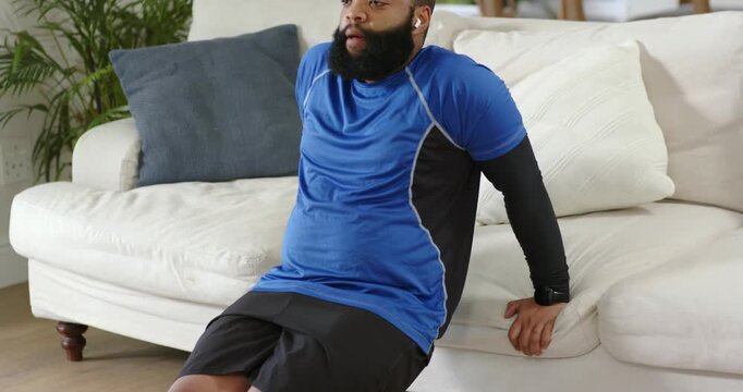 African American man bracing on sofa at home doing dips lowering, rising in shorts for strength