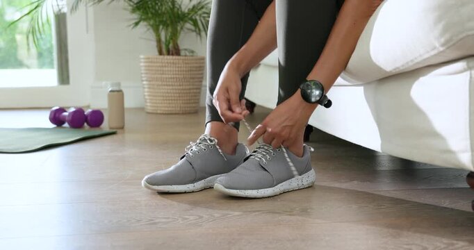 African American female sitting on sofa tying sneaker laces, then checking smartwatch for workout