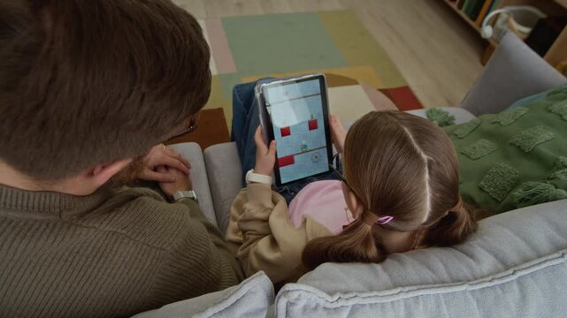 High angle view of father sitting next to young daughter playing game on digital tablet and getting parental control lock screen at home