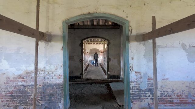 Slow forward tracking shot through an arched doorway in an abandoned, derelict barn, revealing a person standing in the distance.
