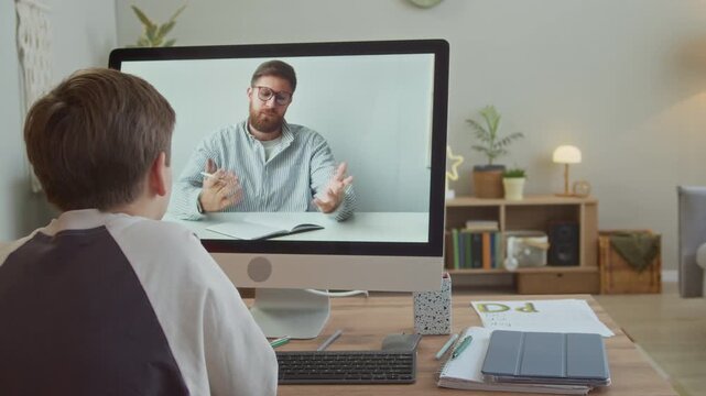 Rear view of young boy listening to male Caucasian tutor during virtual class on computer and finishing lesson from home