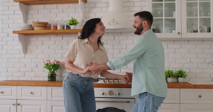 Couple dancing in kitchen at home, laughing and holding hands with joy and love. Young man and woman wearing casual jeans, loving partners smiling, sharing smile and enjoying morning together