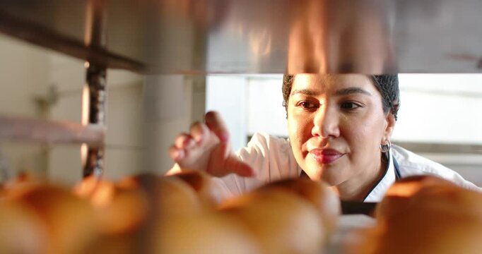 Female baker in chef coat noticing golden rolls, leaning into bakery rack, probing for doneness