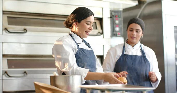 Diverse female bakers in chef coats at prep counter, mentor selecting roll teaching trainee