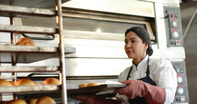 Mid adult female wearing mitts and chef jacket opening oven, pulling tray onto rack, cooling rolls