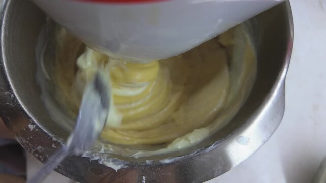 Extreme close-up video of dough mixing in a planetary mixer. A hand holds a tablespoon of soft butter and adds it to the dough. The mixer blends butter into the dough.