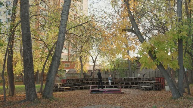 Autumn park empty wooden benches rows under golden canopy of trees, fallen leaves blanket ground, distant brick buildings visible, late afternoon light casts soft shadows, quiet outdoor seating