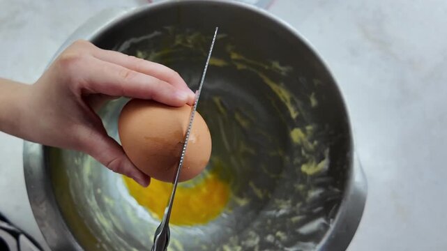 Slow motion close-up of a child's hands cracking an egg with a table knife over a metal bowl. Then separating the shell, egg slowly falls into bowl. Home baking concept, parent-child bonding, learning