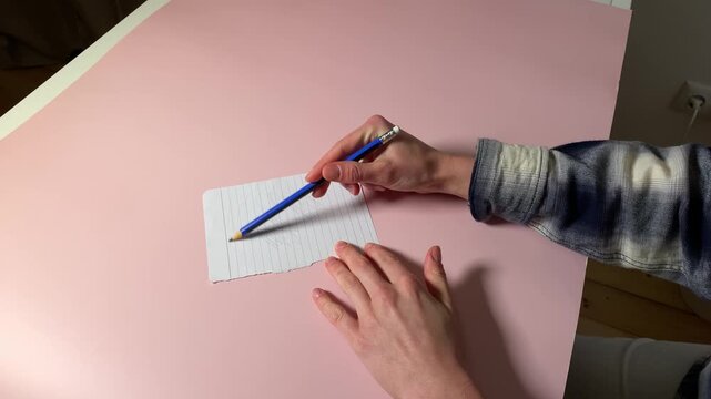Close-up video of a person's hands using a pencil to draw a sketch on a small piece of lined paper. The scene takes place on a clean, pink desk, showing the creative process in motion.