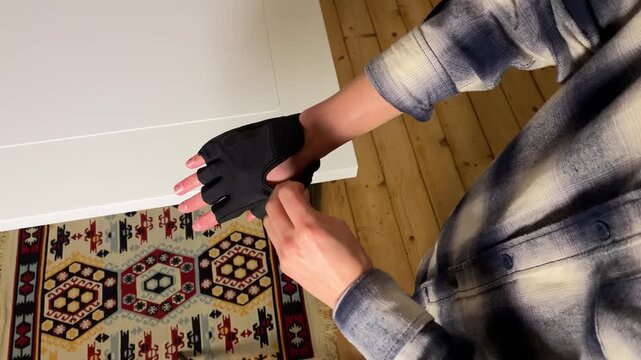 High-angle close-up video of a person putting on and fastening a black fingerless workout glove. Hands get ready for exercise, cycling, or weightlifting over a white table and wooden floor.