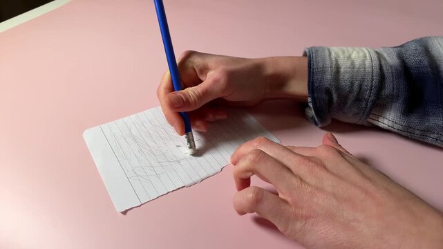Top-down view of a person erasing a pencil sketch on a torn piece of lined paper with a pencil eraser. The scene is set on a soft pink background, suggesting creativity or making corrections.