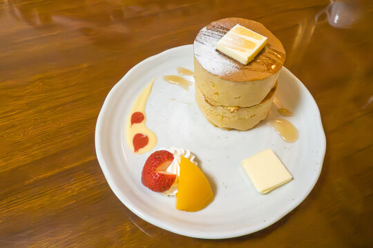 木目調のテーブルに置かれた厚焼きスフレパンケーキの2段重ね。バターとシロップがとろける美味しそうなスイーツに、苺と桃、ホップクリームが添えられたカフェのデザート。左側に広いコピースペースがある広告用素材。