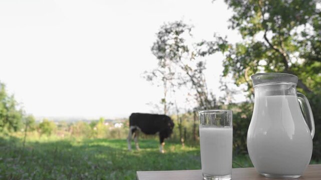 A glass and jug filled with fresh milk are placed on a wooden surface, with a cow grazing in a lush green field behind them. The scene evokes freshness, health, and rural tranquility
