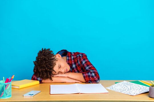 Tired student asleep at desk after studying and doing homework, young boy resting head on arms with open notebook and school supplies on bright blue background