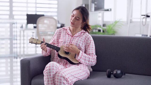 Asian adult woman in pink checkered pajamas playing ukulele sitting on sofa home living room musical instrument practice leisure relaxation hobby