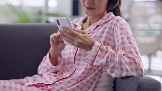 Close up Asian adult woman hands holding smartphone horizontally gaming headphones pajamas sofa home leisure digital entertainment mobile technology