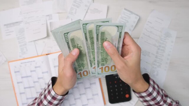 Close up of male hands holding and counting US dollar bills, surrounded by paperwork and a calculator. The scene represents personal finance management, accounting, and expense tracking