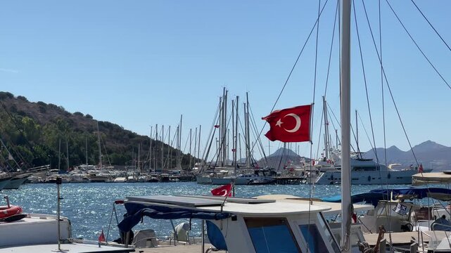 A vibrant Turkish flag flutters in the breeze at a sun-drenched marina in Bitez, Bodrum. White yachts and sailboats dot the sparkling blue Mediterranean harbor under a clear sky.