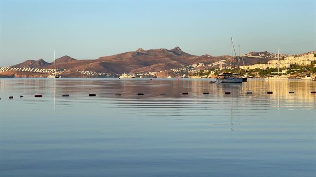 A tranquil sunrise over Bitez Bay, Bodrum. Sailboats rest on glassy, mirror-like water, reflecting the golden light and coastal hills of the Turkish Riviera in a serene, panoramic view.