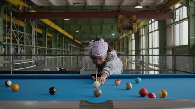 Front view medium shot of young Biracial man wearing hipster clothes aiming with cue and striking cue ball during fun billiards game in industrial loft setting