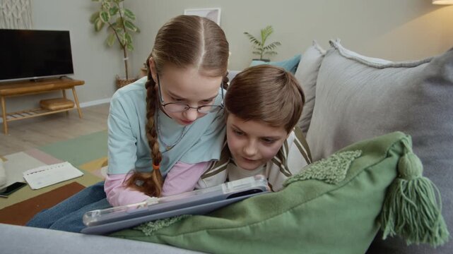 Handheld shot of young Caucasian kids resting on sofa while playing on digital tablet together in cozy living room