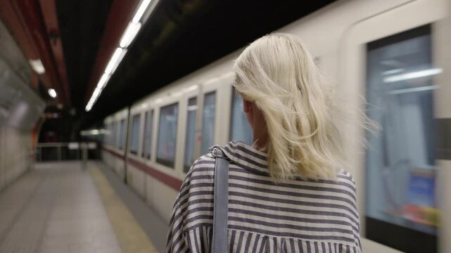 Young woman standing on platform with wind blowing hair from approaching train, urban transport atmosphere