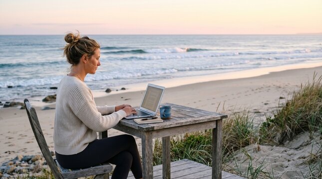 Young woman typing on laptop at a rustic wooden table by the ocean during sunset for remote work concept and digital nomad lifestyle