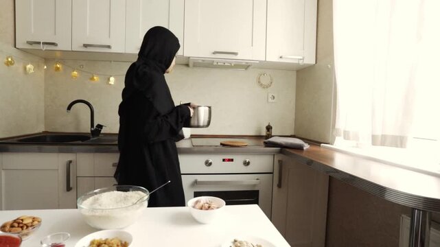 muslim woman in black hijab carrying a metal cooking pot in a bright kitchen during home meal preparation for ramadan, iftar or festive family dining
