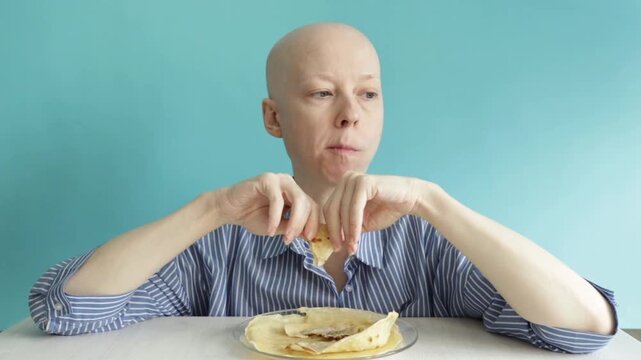 bald female oncology patient eats pancakes,chews slowly and listens to food sounds,home meal routine during chemotherapy recovery with calm blue background and copy space