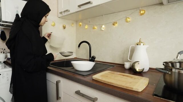 wide shot of a woman in hijab draining rice into a strainer over a white bowl in a bright home kitchen, documenting ingredient preparation for a festive family dish