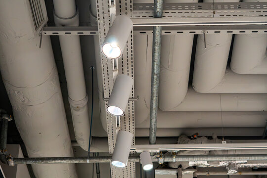 Modern track lighting and white LED spotlights on an industrial ceiling, visible ventilation pipes and technical installations in loft style, interior lighting for commercial and office spaces.