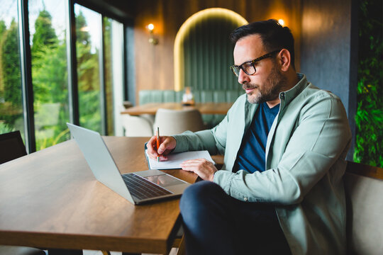 Focused businessman writing notes while working on laptop in cafe