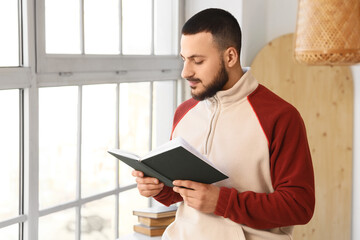 Handsome young man reading book near window at home