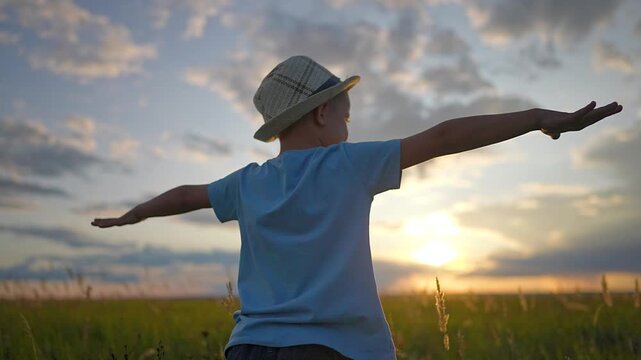 Little boy stands in green field at sunset with open arms. Happy child dreams of flying like airplane. Boy in hat enjoys nature freedom. Kid plays pilot in summer sunset field. Boy looks at sky.