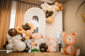 Beautifully decorated photo zone for a child's birthday party. Features a white arch, balloons in cream and brown tones, and large cutouts of a bear, bunny, and mushrooms in a forest theme. © Serhii