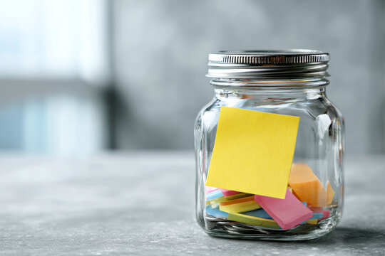 Glass jar filled with colorful sticky notes on a gray table, with a yellow note attached to the front. Creative organization and productivity concept for modern workspaces and brainstorming sessions
