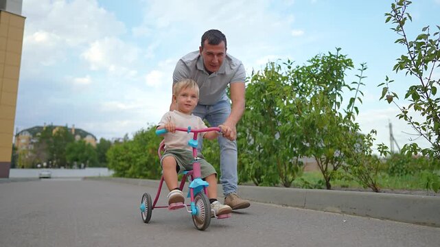 Father pushes little son on tricycle on paved path. Happy man helps boy ride bike near building. Family enjoys summer walk outdoors. Dad teaches kid to ride three-wheeled bike during day.